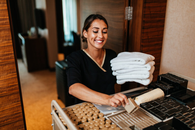 Beautiful young hotel housekeeper in uniform bringing clean towels and other supplies to hotel room.